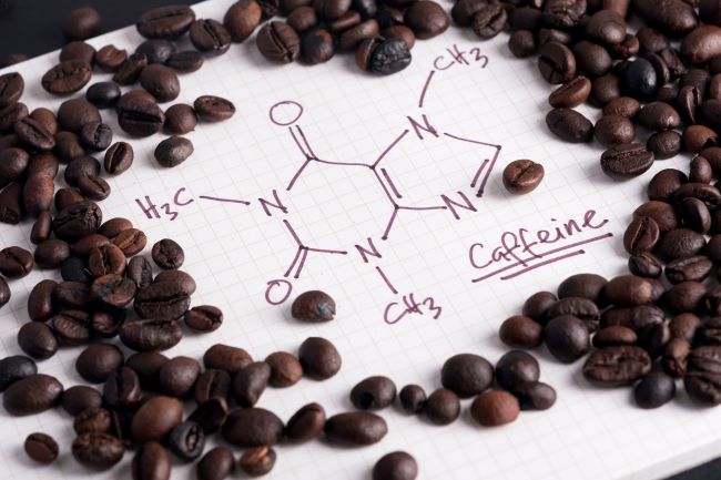 Na kostkovaném papíře je napsán chemický vzorec kofeinu, kolem jsou hnědá kávová zrna.